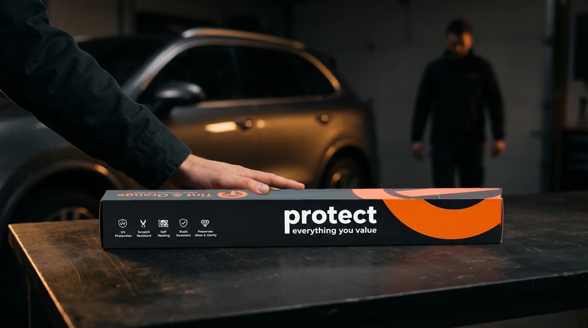 A hand resting on a sealed Tint & Orange Interior Protection Film carton in a garage, with the installer waiting in the background